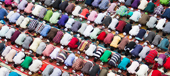 A huge crowd of Muslims praying around the Kaaba in Mecca with open, praying hands in the foreground. 